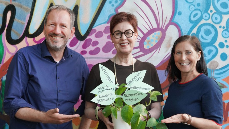 Andreas Gleis, Birgit Westers und Alexandra Heckhuis mit einer Pflanze, deren Blätter mit Begriffen zur Mitbestimmung von Kindern beschriftet sind.