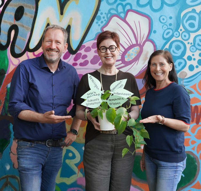 Andreas Gleis, Birgit Westers und Alexandra Heckhuis mit einer Pflanze, deren Blätter mit Begriffen zur Mitbestimmung von Kindern beschriftet sind.